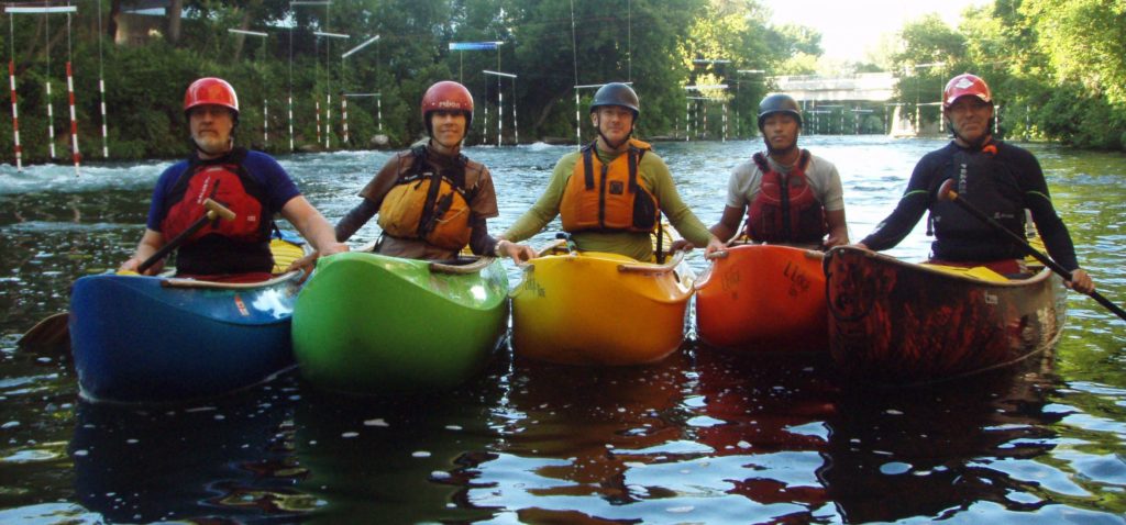 Five canoeists in helmets and life-jackets side-by-side in multi-coloured canoes